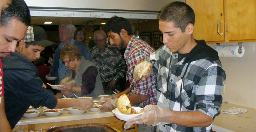 young college-age men dish up bowls of beans from a large slow cooker.